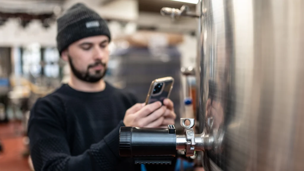 PLAATO-Sensor an einem Edelstahl-Fermentationstank mit Person im Hintergrund, die Messdaten per Smartphone überprüft